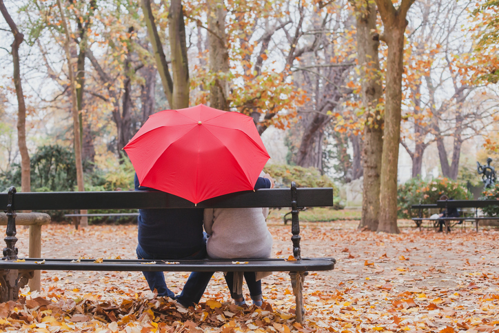 Rear view of couple seated on park bench, in close contact, red umbrella hiding their heads from view