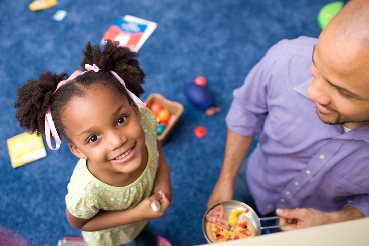A little girl in a playroom smiles up at the camera while parent smiles down at her
