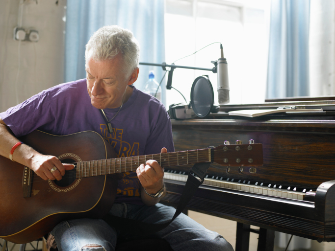 Mature man plays a guitar while seated at piano bench