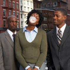 A woman in a business suit blows a bubble with her gum while two businessmen look at her with odd expressions.