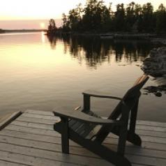 An empty wooded chair sits on a wooden dock as the sun sets over a lake.