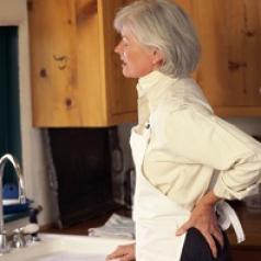 A woman stands at kitchen sink in obvious back pain. 