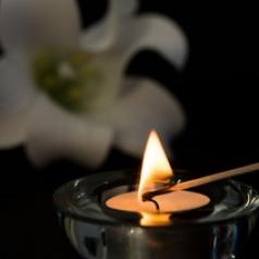 A small votive candle is being light with a match, and a white flower lies in the background.