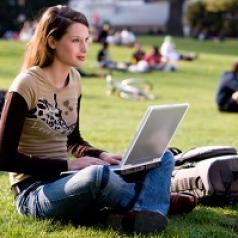 A young woman sits on the grass with her laptop on a college campus.