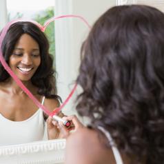 Young adult with curly long hair draws heart on mirror with lipstick, smiling