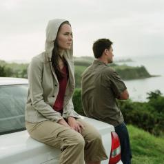 Couple looks at coastline, man stands, woman sits on back of car