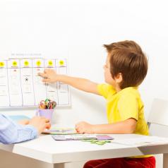 Young boy sits with parent at small table and adds activity to calendar on wall