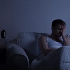 Person sits on sofa in dark room, covering eyes with one hand