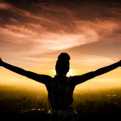 Woman faces sunset and raises arms in triumph.