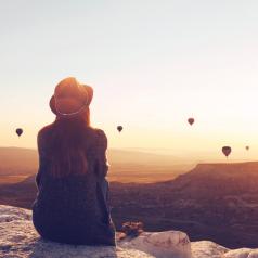 A young woman sits and watches hot air balloons drifting across the horizon.