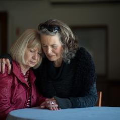 Two senior women hug while sitting at a small library table.