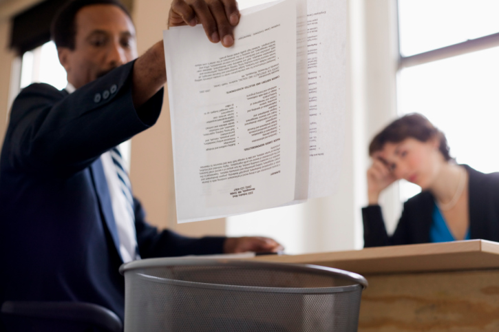 Businessman tosses report into wastebasket while woman watches in annoyance