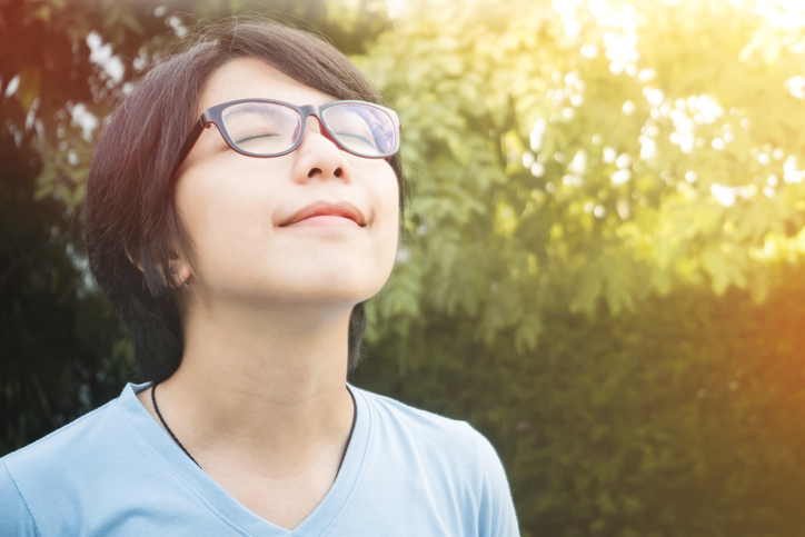 young person wearing glasses looks up into the sky and breathes deeply, smiling, eyes closed