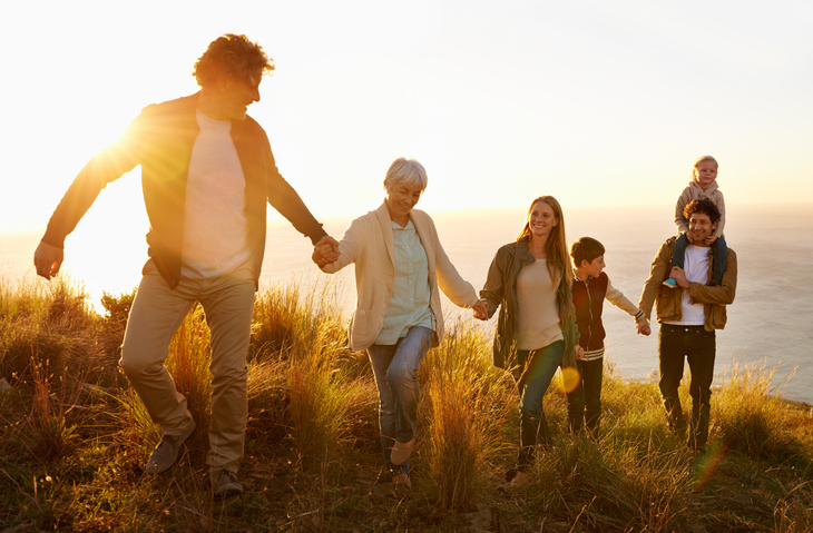 Three generations of family members hold hands and walk in a line across hilly field at sunset