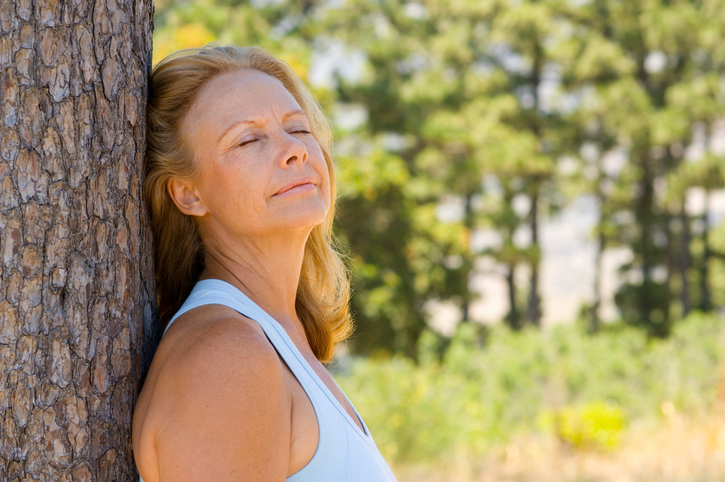 Calm, tanned person with long golden hair rests haid against tree, eyes closed