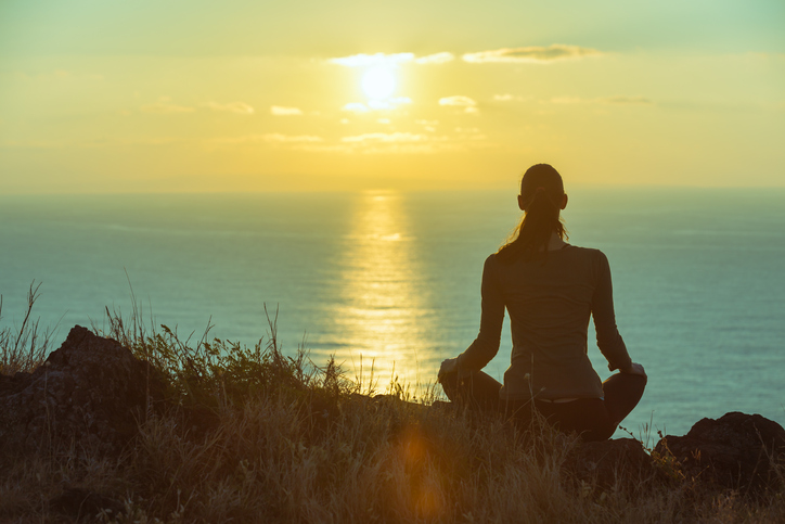 Adult with ponytail sits on hill in athletic wear in meditative pose, watching the sun rise