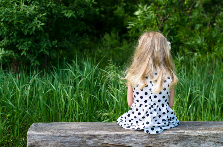 Rear view of a child with long blonde hair sitting on dock facing grassy overgrowth