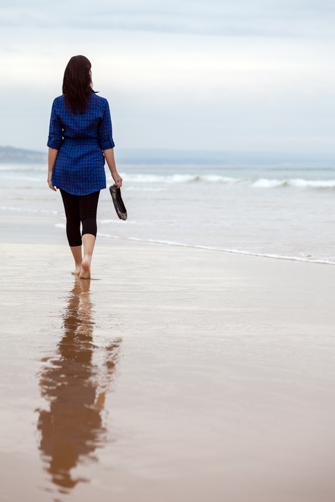 Rear view of person wading in shallow beach, shoes in right hand