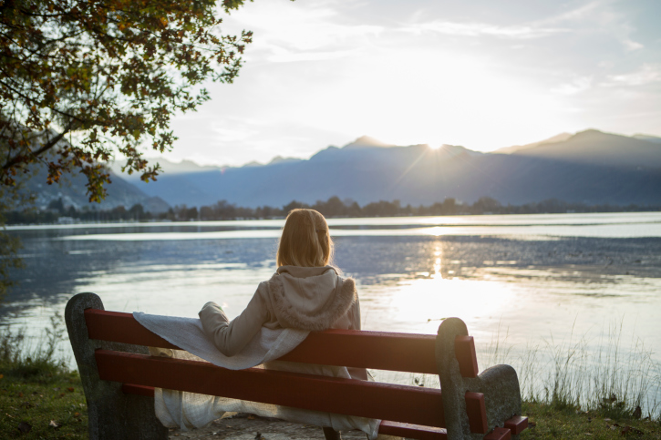 A woman sits on a bench under a tree and looks out at a lake