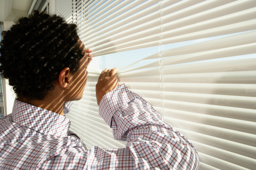 A young man in an office peers through blinds