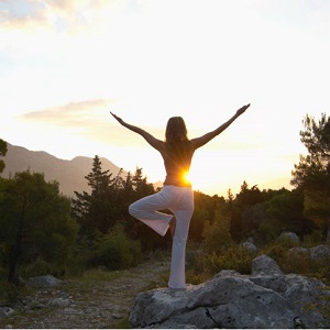 A woman does a yoga pose in a natural setting with the sun rising in the background.