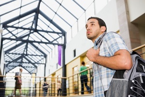 Nervous-looking young man with short dark hair, wearing plaid shirt and backpack. He is standing in a glass-ceilinged college building with many people walking behind him.