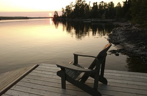 An empty wooded chair sits on a wooden dock as the sun sets over a lake.