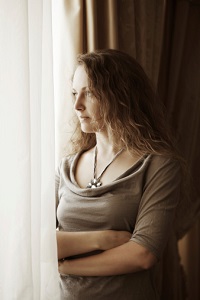 A woman stands hugging herself, looking out a window.