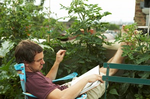 A man, his feet kicked up on a bacolny, reads a book near plants.