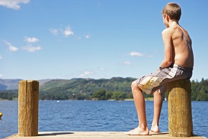 A shirtless boy sits on a dock, looking out over water.