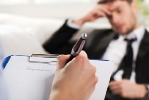 A therapist writes "depression" on a paper attached to a clipboard while a client sits in front of him with his hand on his head.