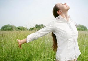A woman stands in a grassy field and streatches her arms out while taking a deep breath.