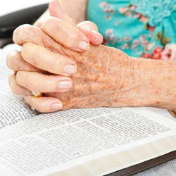 An elderly woman's hands are folded in prayer while resting on a bible.