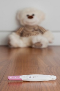 Out of focus a stuffed bear leans against a wall behind a pregnancy test stick, in focus. 
