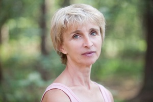 A woman stands in woods, looking at a camera. 