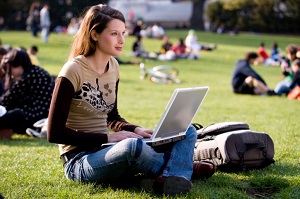 A young woman sits on the grass with her laptop on a college campus.