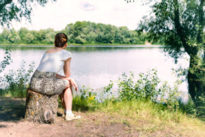 Person in skirt with hair pulled back sits on tree stump at lakeside looking over water