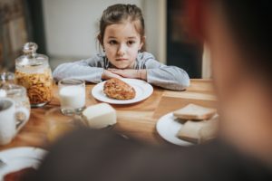 Young girl looks unhappy as she sits at the table with her parent at breakfast