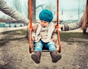 Toddler sitting on orange swing, with arms of both parents in frame swinging her forward