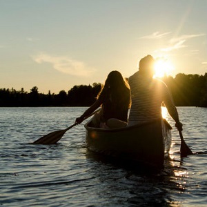 A couple paddles in a canoe on a lake, heading toward a bright sunset.