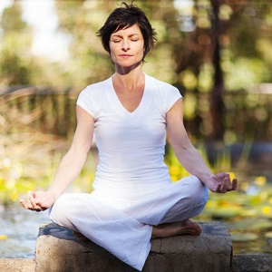 A woman sits outside in a peaceful area and meditates.