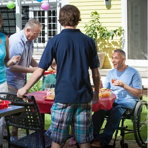 A man in a wheel-chair smiles and enjoys a barbeque with his family.