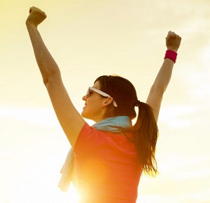 A woman raises her arms in victory as she stands outside facing the sun.