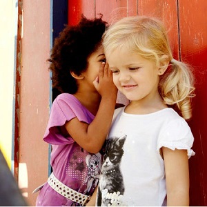 One girl whispers to another while standing outside in a hidden place.