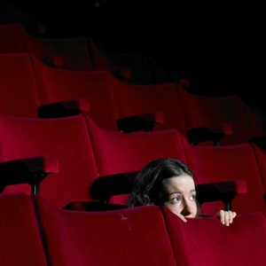 A young woman crouches down and peers over a seat in an empty theater.