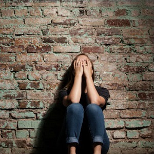 A woman crouches against a brick wall and covers her face.
