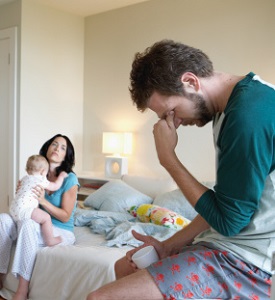Young white couple wearing pajamas and sitting in their messy bedroom. The mother is sitting on the bed, looking exhausted and angry, holding her baby in her lap. The father is sitting across the room, holding a cup of coffee with his face in his hands.