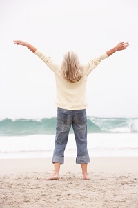 A woman faces the ocean with her arms in the air, in a joyful posture.