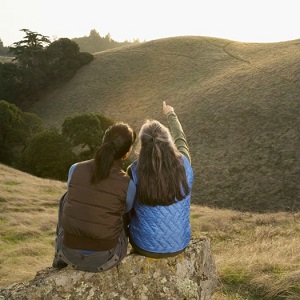 Two women sit on a rock looking out at a hillside.