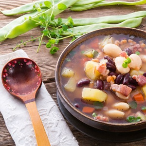 A colorful wooden spoon sits next to a stew with many ingredients; snap-peas and fresh herbs surround the bowl.